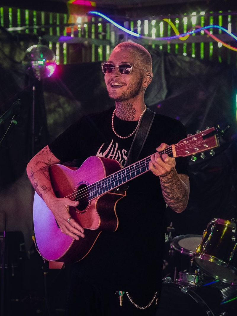 Joshy playing an acoustic guitar on stage, lit in deep magenta, with a drum kit and green stage lights behind.