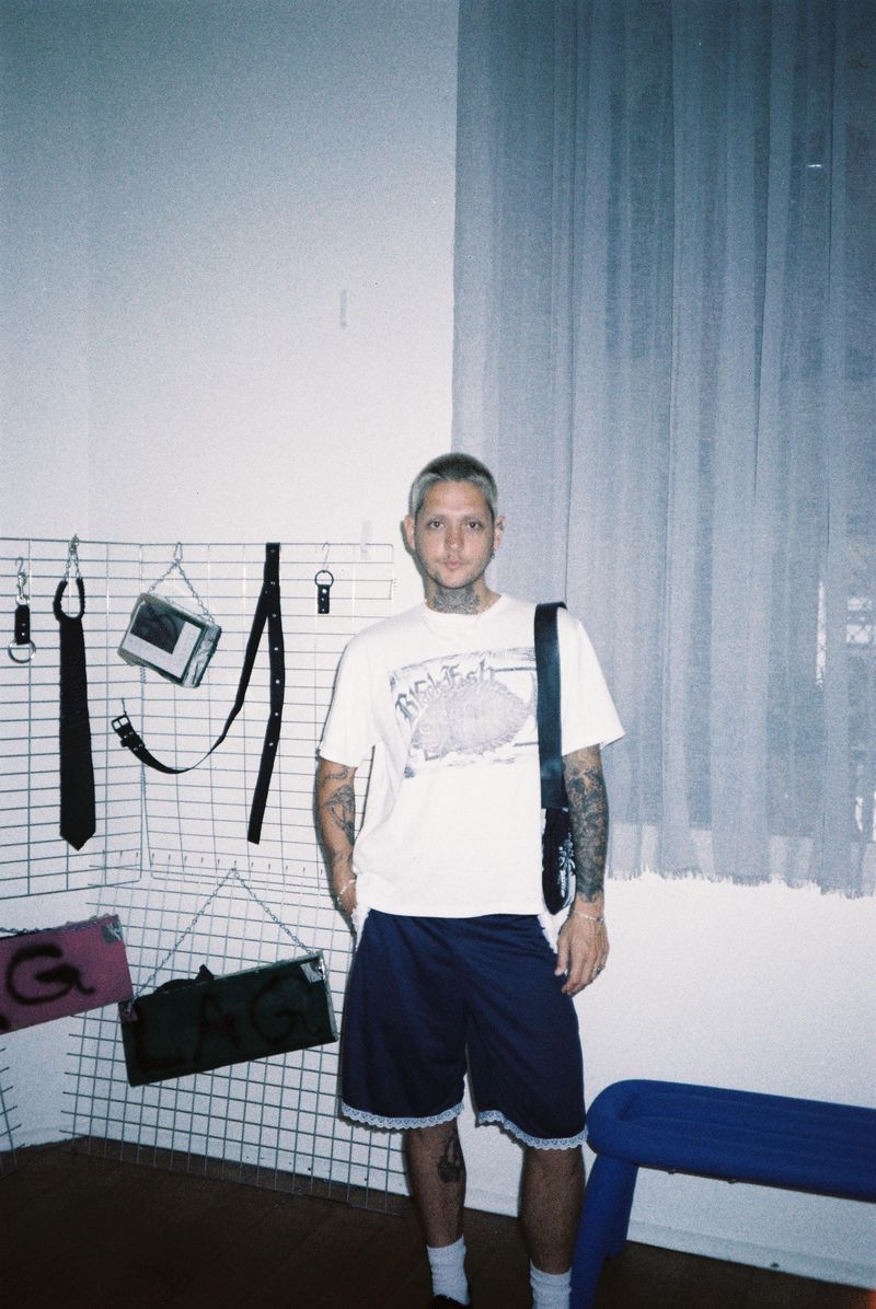 Joshy standing in a white-tiled room in front of a pegboard hung with tools, wearing a printed t-shirt and shorts.