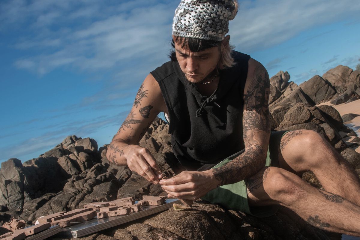 Joshy crouched barefoot on dark coastal rocks under an open blue sky, working a piece of clay in both hands with tools laid out on the stone beside them.