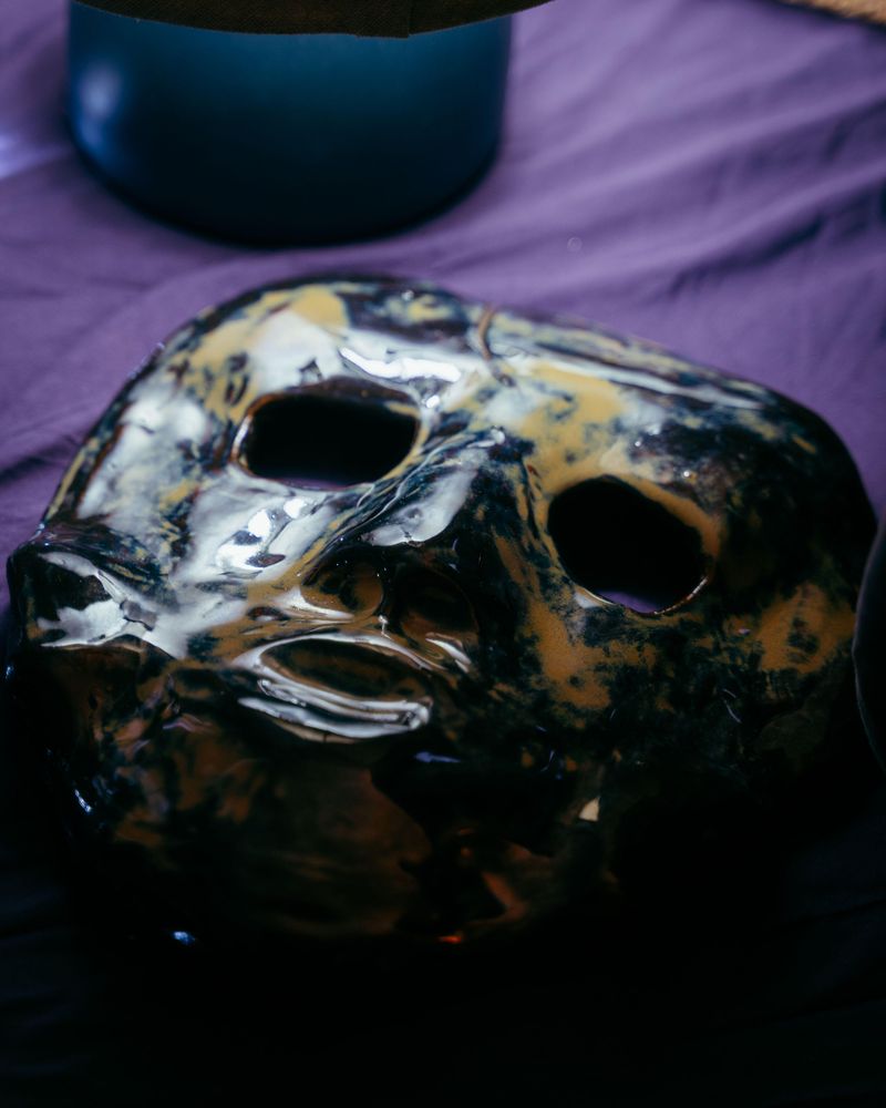 A hand-formed ceramic mask lying on purple cloth, its thick glaze pooling in glossy black, amber and cream across the brow and cheeks. The eye openings and mouth are cut irregularly. A dark metal bowl sits behind it.