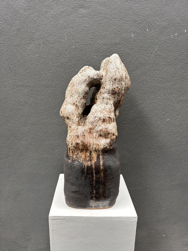 The same sculpture after firing, its surface now pale and scorched with dark iron blooms, on a white shelf against a grey wall.
