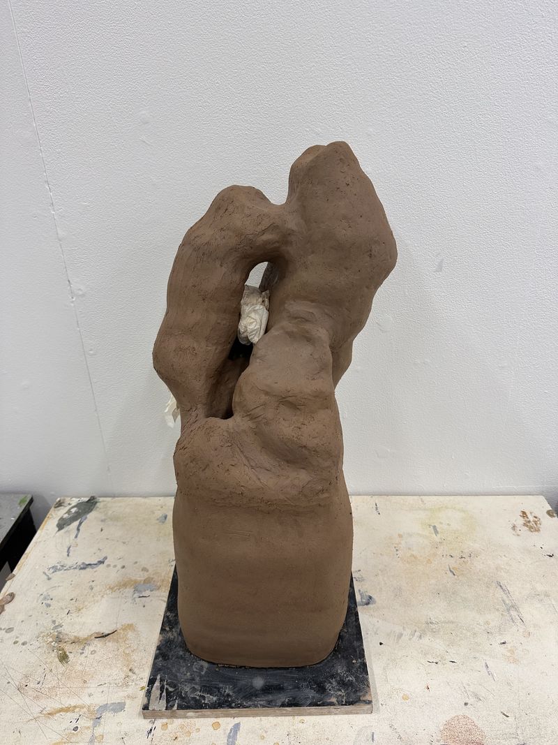 An unfired clay sculpture, raw brown and hand-formed into a curling organic shape, mounted on a white shelf.
