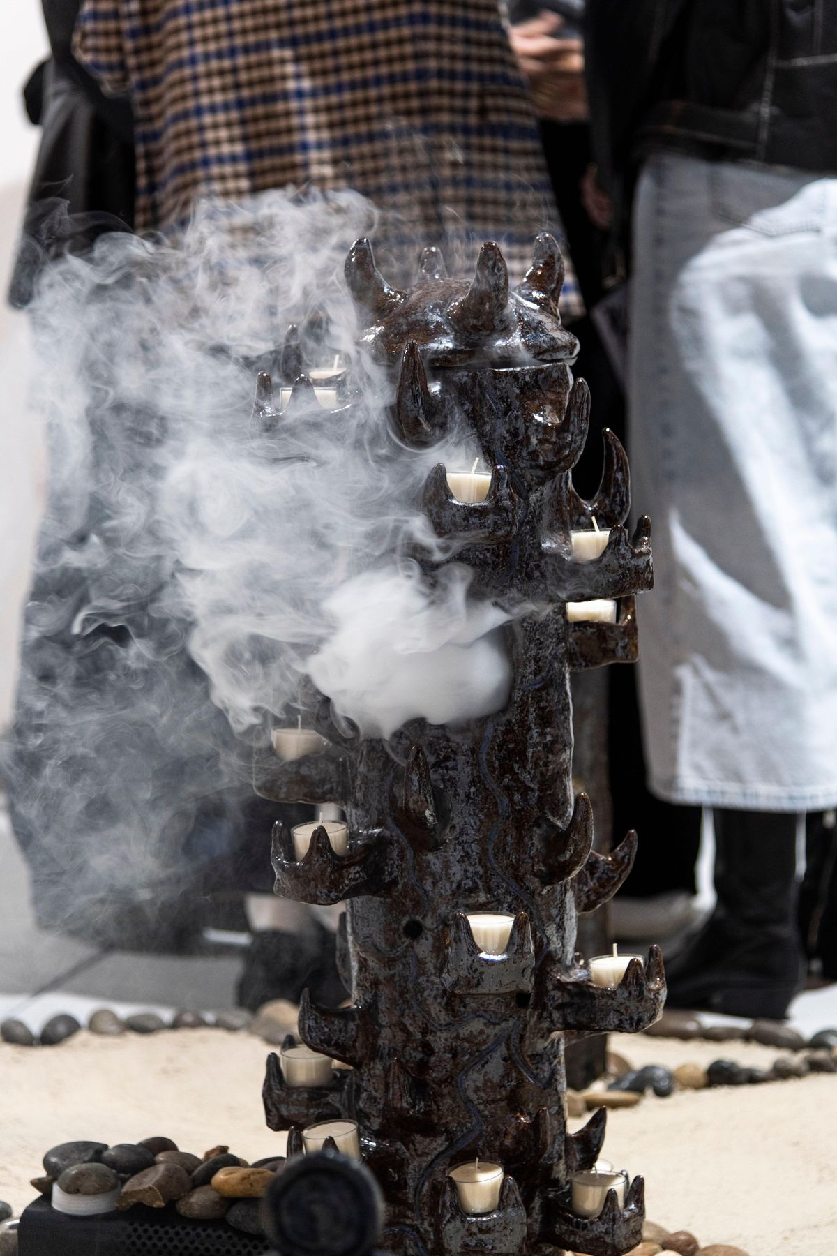 A tall dark ceramic column studded with small candles, standing in a ring of stones as smoke rises around it.