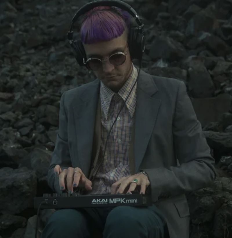 Joshy in a tailored suit and over-ear headphones, playing a small MIDI keyboard while seated on a bed of grey stones.