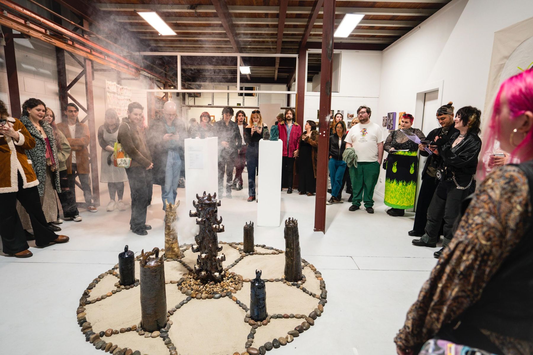 A warehouse gallery where seven dark ceramic works are arranged in a circle of stones on the floor around a central plinth, with an audience standing in a ring watching.