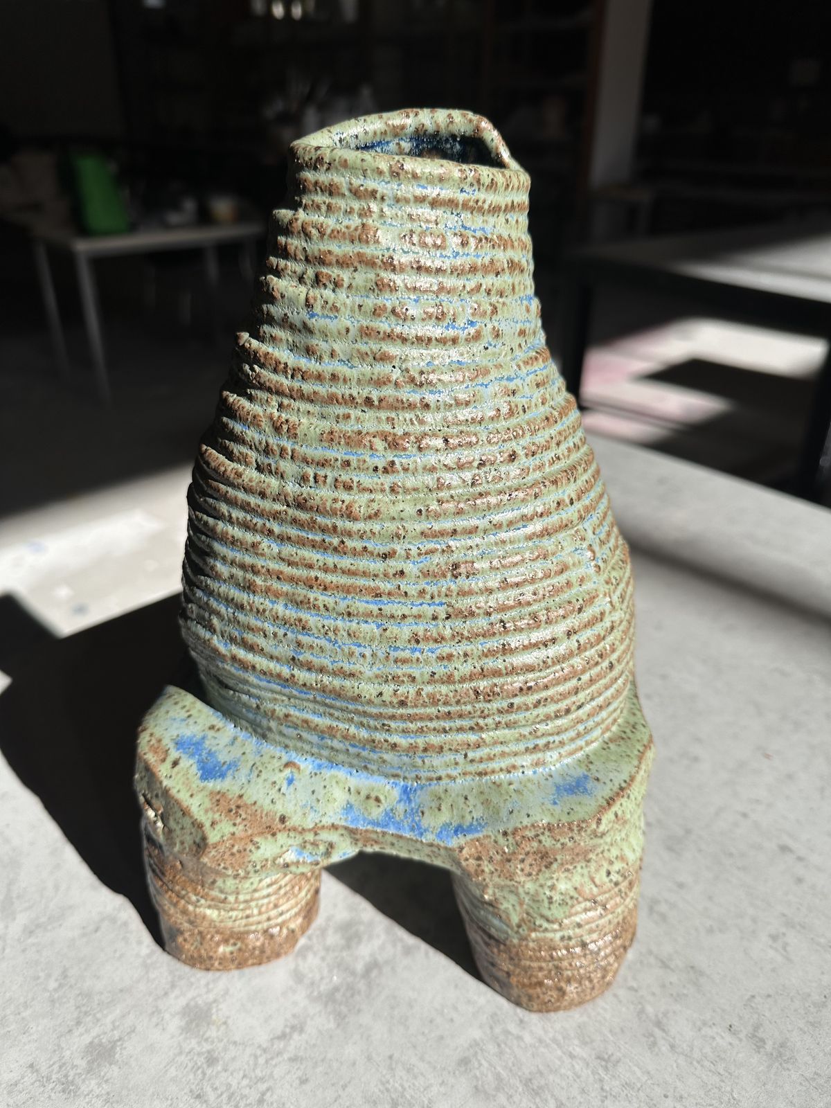 A coil-built ceramic vessel with deeply ridged walls and a pale green-blue glaze, lit by hard direct sunlight.
