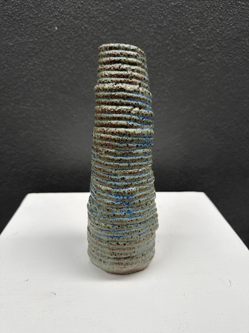 A ridged, tapering ceramic form with a mottled green and pale blue surface, resting on a white gallery plinth against a dark wall.