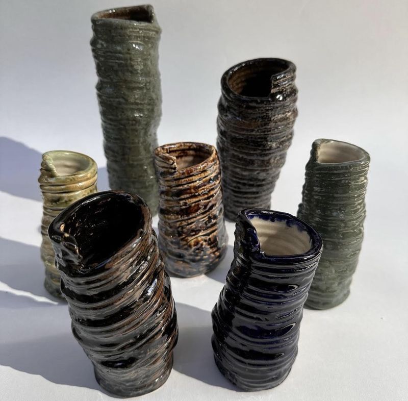 Seven ridged ceramic vessels of varying height grouped on a pale surface, casting long hard shadows. Glazes range from near-black gloss to olive and pooled amber.